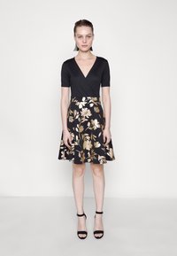 Robe noire à manches courtes, décolleté en V et jupe évasée avec des motifs floraux dorés. Le mannequin porte des talons noirs avec une bride à la cheville.