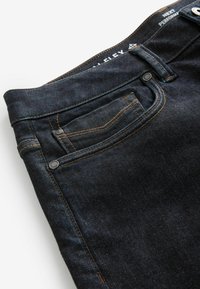 Jeans de mezclilla oscura con costuras visibles, un pequeño bolsillo frontal y una textura suave. Presenta un cierre de botón y cremallera metálica.