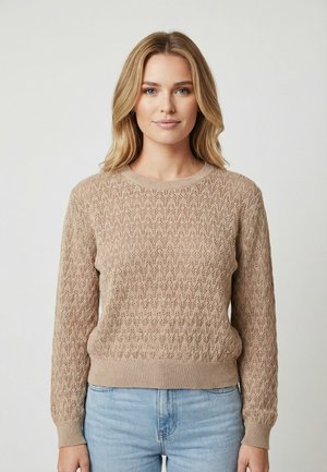 Donna bionda che indossa un maglione lavorato a maglia di colore marrone chiaro e jeans azzurri, in piedi davanti a uno sfondo bianco uniforme.