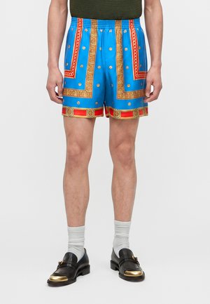 Άνδρας μοντέλο φοράει μπλε σορτς με χρυσά και κόκκινα μπαρόκ μοτίβα, συνδυασμένα με λευκές κάλτσες και μαύρα loafers με χρυσές μύτες.