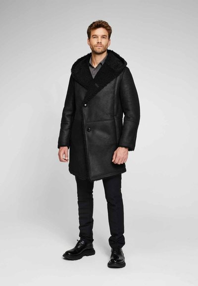 Cappotto di pelle nera con un ampio colletto peloso, chiusura frontale con bottoni e un design dritto; abbinato a pantaloni neri e scarpe.