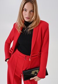 Rode op maat gemaakte blazer met een gestructureerd ontwerp, gecombineerd met bijpassende broeken. Zwarte geribbelde coltrui eronder, met een glanzende zwarte clutch in de hand.