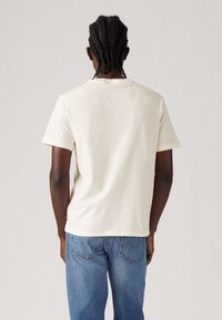 Lys beige bomull t-skjorte med rund hals og korte ermer, vist fra baksiden. Blå denimjeans med lommer.