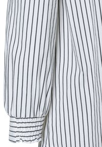 White shirt with vertical black stripes, featuring a gathered elastic cuff and soft, lightweight fabric. Striped pattern runs throughout.