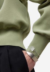 Sweter o oliwkowozielonej fakturze z prążkowanymi mankietami, zestawiony z czarnym ubraniem. Srebrna bransoletka z przezroczystymi zdobieniami na nadgarstku.