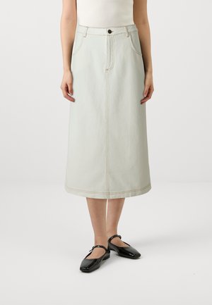 Femme portant une jupe midi beige clair taille haute avec poches avant et des chaussures plates Mary Jane noires, debout sur un fond uni.