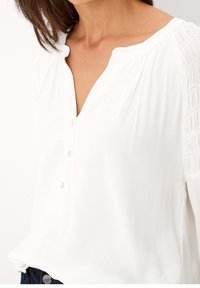 Blusa bianca realizzata in tessuto morbido con scollatura a V, dettagli con bottoni e tessuto arricciato sulle maniche. Il design è leggero e rilassato.
