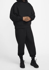 Černá mikina s kapucí a matching tepláky, vybavené přední kapsou, žebrovanými manžetami a texturovanou látkou. Nošeno s černými teniskami.