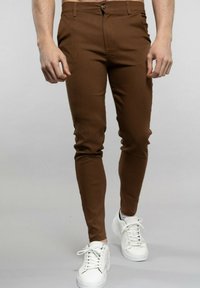 Pantalons slim marron en tissu lisse, avec une fermeture à bouton, des poches latérales, portés avec des baskets blanches.