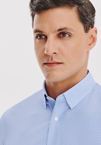 Mężczyzna z krótkimi ciemnymi włosami, ubrany w jasno niebieską koszulę w kratkę z guzikami, patrzy lekko w prawo na białym tle.