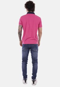 Homme tatoué portant un polo rose, un jean bleu et des baskets blanches, debout de dos sur un fond uni.