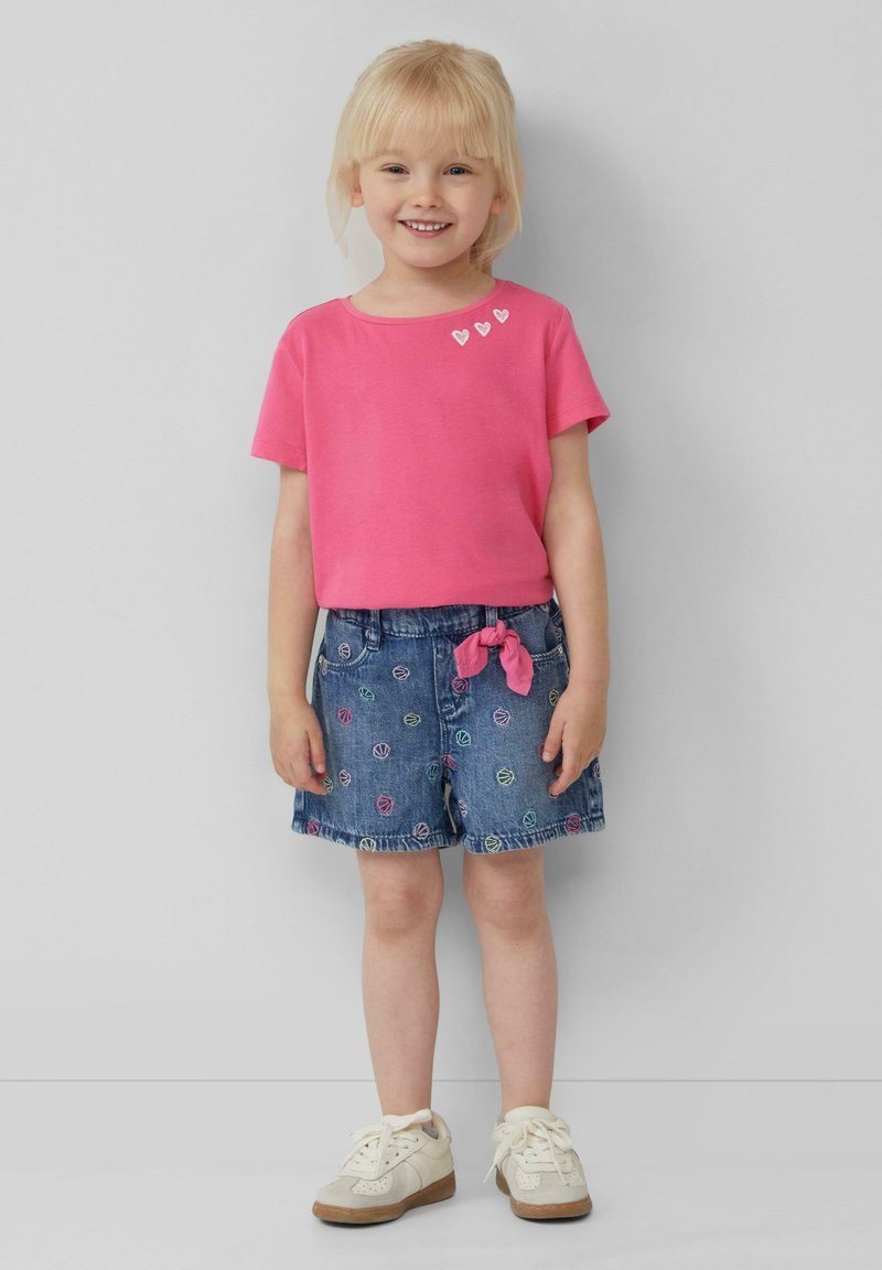 Roze shirt met korte mouwen met drie kleine witte hartpatronen; denim shorts met meerkleurige balmotieven en een roze strik in de taille; witte sneakers.