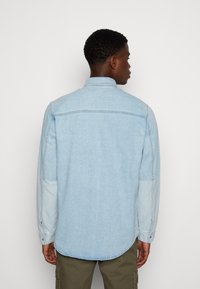 Camicia di jeans azzurro chiaro con maniche lunghe, colletto appuntito e orlo curvo. Presenta pannelli delle maniche a contrasto e polsini con bottoni.