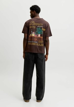 Homme portant un T-shirt marron foncé avec un motif coloré de plantes et d’objets et le texte "Perfect Days 24/7 House Rules" au dos, associé à un jean noir ample.