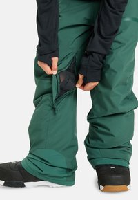 Groene waterdichte sneeuwbroek met ritsventilatie en een mesh voering, gecombineerd met zwarte snowboardlaarzen met een gestructureerd ontwerp.