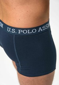 Marineblå boksershorts laget av et glatt, elastisk materiale med en kontrastfarget logo på midjebåndet. Sømmer synlige langs sidene.