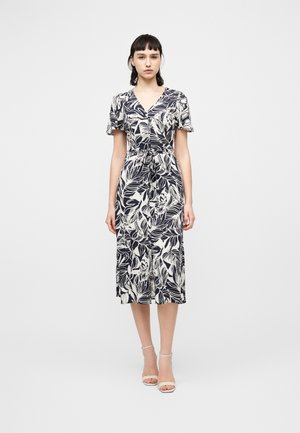 Γυναίκα που στέκεται φορώντας μια μεσαίου μήκους φόρεμα με μαύρο και άσπρο φυλλώδες μοτίβο, κοντά μανίκια και λευκές γόβες με λουράκι στον αστράγαλο σε απλό φόντο.