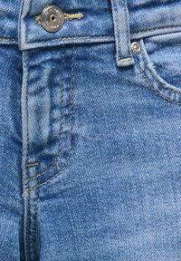 Gros plan sur un jean en denim bleu montrant un bouton en métal, une passants de ceinture et les détails des coutures de la poche avant.