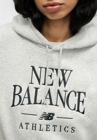 Ανοιχτό γκρι φούτερ με κουκούλα που διαθέτει μαύρο κεντητό κείμενο "NEW BALANCE ATHLETICS", ρυθμιζόμενο κορδόνι και απαλό ύφασμα.