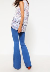 Top senza maniche con motivo floreale in colori chiari abbinato a jeans a zampa d'elefante blu. L'outfit presenta una vestibilità rilassata e una texture morbida.
