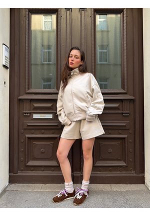 Νεαρή γυναίκα με μπεζ μπουφάν και σορτς με καφέ αθλητικά παπούτσια στέκεται μπροστά σε μια μεγάλη σκούρα καφέ ξύλινη πόρτα με γυάλινες πλάκες.