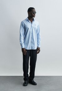 Άνδρας που φοράει ριγέ ανοιχτό μπλε πουκάμισο με κουμπιά, μαύρο παντελόνι, μαύρα παπούτσια και γυαλιά ηλίου, στέκεται μπροστά σε απλό φωτεινό φόντο.