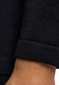 Dunkel gestrickter Stoff mit erhabener Textur und umgeschlagenem Bündchen. Nahaufnahme zeigt glatte Nähte und den kontrastierenden Hautton des Handgelenks.