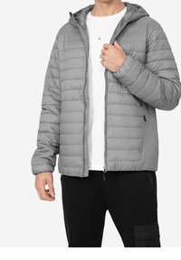 Doudoune grise avec coutures horizontales, fermeture éclair, capuche attachée et poignets élastiques, portée sur une chemise blanche et un pantalon noir.