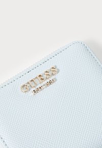 Gaiši zils teksturēts maks ar zelta "GUESS EST 1981" logotipu, ar rāvējslēdzēju un smalkiem šuvuma akcentiem pa malu.