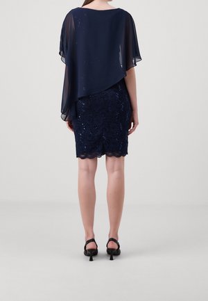 Donna che indossa un abito in pizzo blu navy con paillettes, con un soprabito asimmetrico trasparente e sandali neri con tacco, vista di spalle su uno sfondo semplice.