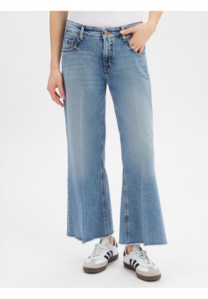 Person trägt hellblaue, weit geschnittene, gekürzte Jeans mit rohen Säumen und weiße Sneakers mit schwarzen Streifen.