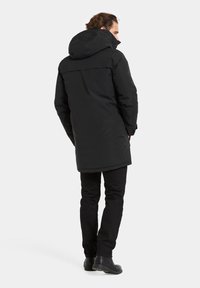 Parka d'hiver noire avec capuche, présentant un design mi-long, une texture lisse et des coutures minimales. Portée par-dessus un pantalon noir et des bottes.