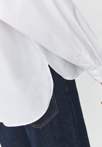 White long-sleeve shirt with a curved hemline, paired with dark blue denim jeans featuring contrasting orange stitching and a side pocket.
