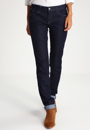 Mujer vestida con jeans azul oscuro ajustados de corte slim, con los puños doblados, combinados con botines de ante marrones y una blusa blanca.