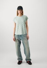 Top de manga corta verde claro con patrón, combinado con jeans de corte holgado azul claro y una gorra negra. Los zapatos presentan detalles en negro y rojo.