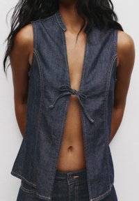 Gilet en denim bleu foncé, présentant un design sans col, une fermeture devant par lien et des détails de couture visibles. Sans manches, avec une coupe décontractée.