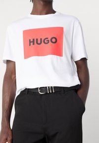 Camiseta de algodón blanca con un gran gráfico rectangular rojo que presenta la palabra "HUGO" en texto negro en negrita. Combinada con pantalones negros y un cinturón.