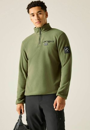 Young man wearing olive green quarter-zip fleece and black cargo pants holding a black bag against a plain background.
