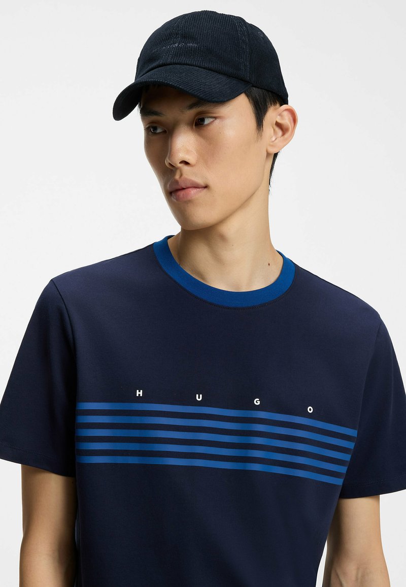 Jeune homme portant une casquette noire et un t-shirt bleu marine à rayures bleues avec le mot HUGO imprimé sur la poitrine, regardant sur le côté.