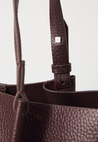 Sac à main en cuir bordeaux avec une finition texturée. Doté d'une bandoulière robuste avec un accent en métal rectangulaire et un détail de boucle réglable.