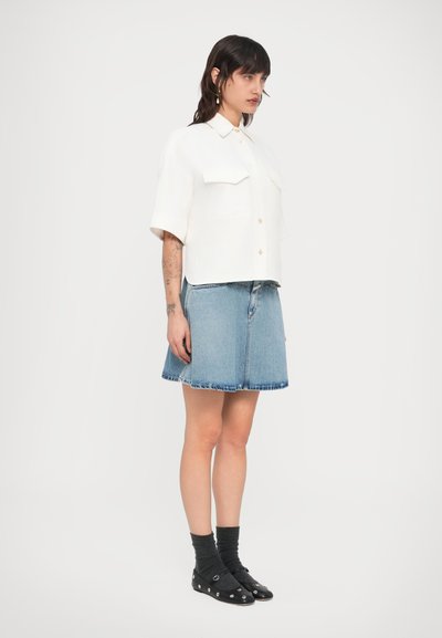 Κοντοκομμένο, λευκό πουκάμισο με κουμπιά και κοντά μανίκια, με δύο τσέπες στο στήθος, σε συνδυασμό με μία μπλε φούστα από τζιν. Μαύρα παπούτσια με διακοσμητικές λεπτομέρειες.