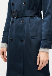 Ναυτικό διπλόστροφο παλτό μπουφάν με μαύρα κουμπιά, ζώνη στη μέση, πλαϊνή τσέπη και ορατά μανίκια γκρι πλεκτού πουλόβερ, φορεμένο από ένα άτομο.