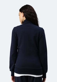 Felpa navy con maniche lunghe e colletto alto, realizzata in materiale morbido. Presenta polsini e orlo a costine, indossata sopra un indumento bianco.