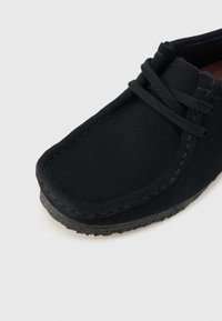 Μαύρο μοκασίνι με σουέντ υφή, με λεπτομέρειες ραφών και κορδόνια δεμένα πάνω σε σόλα από ιξώδες καουτσούκ.