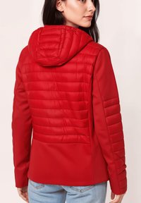 Mujer vestida con una chaqueta acolchada roja con capucha, vista desde atrás, combinada con unos jeans de color azul claro, sobre un fondo liso.