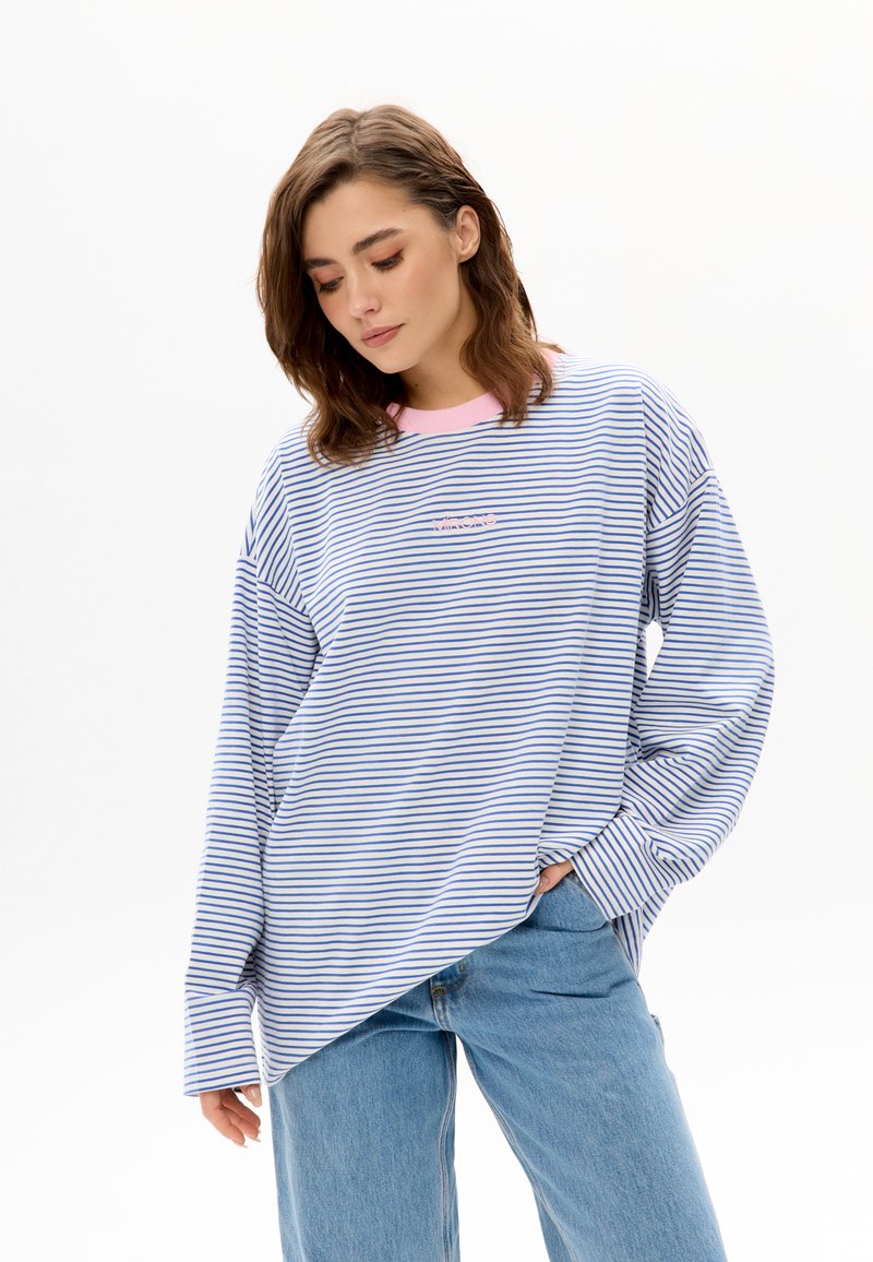 Blauw-wit gestreept shirt met lange mouwen en roze kraagaccenten. Losse pasvorm, zijsplitsen en een klein logo op de borst. Draagt lichtgekleurde spijkerbroek.
