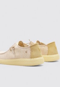 Zapatillas de tela beige con acentos amarillo claro, punta redonda, textura suave, diseño bicolor y suela de goma elevada. Cordones y detalle del logo presentes.