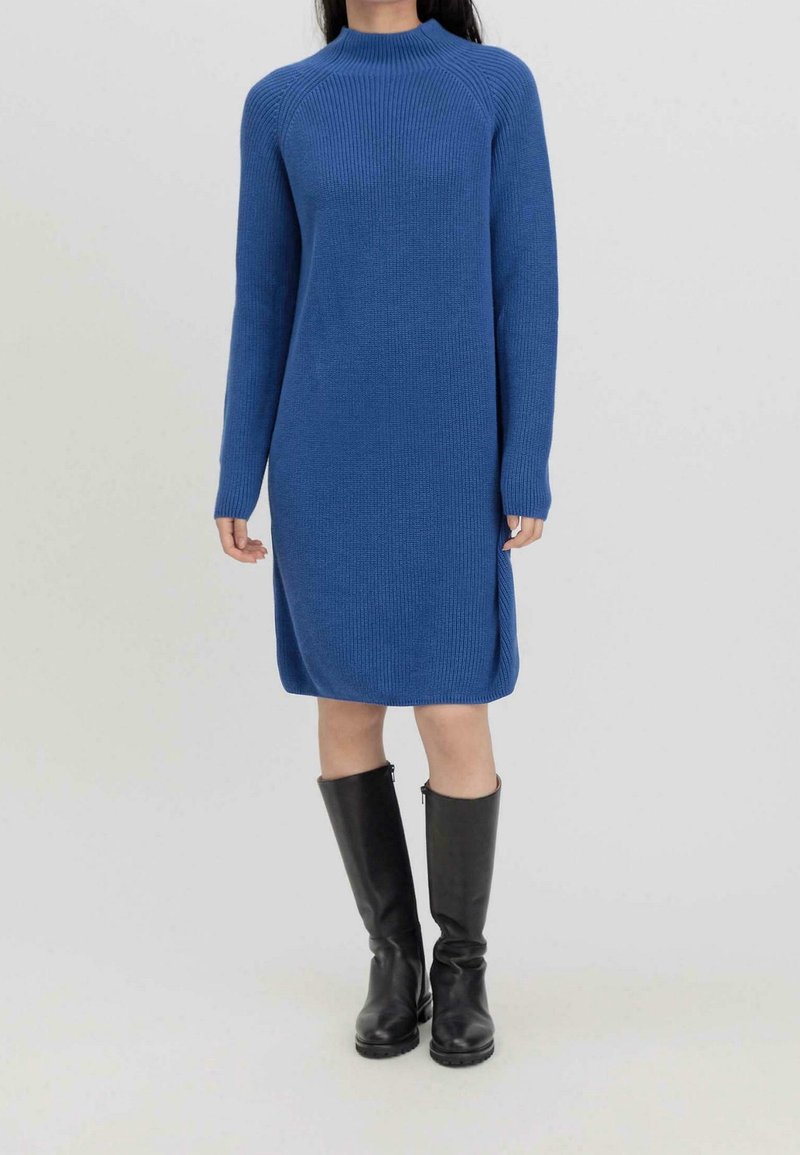 Blå stickad klänning med långa ärmar, hög krage, sidoflikar och ribbad textur, i kombination med knähöga svarta stövlar.