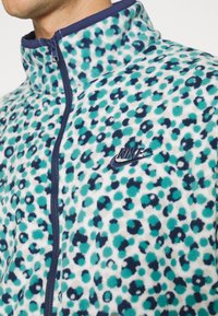 Jachetă din fleece cu un model într-o combinație de pete albastre și verzi, fermoar frontal, guler înalt și logo-ul Nike brodat pe piept.