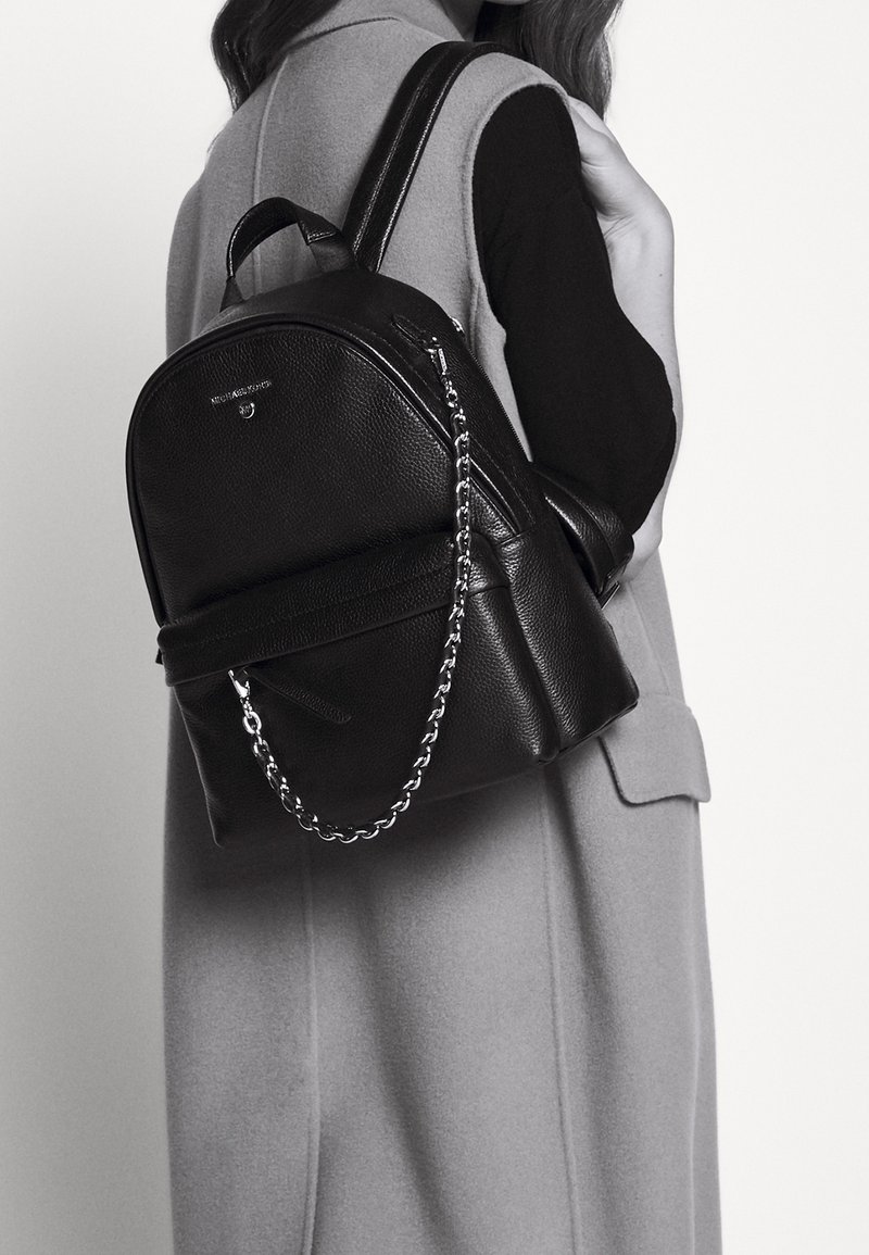 Femme portant un sac à dos en cuir noir avec un détail en chaîne argentée sur un manteau gris sans manches et un haut noir à manches longues, vue de côté.